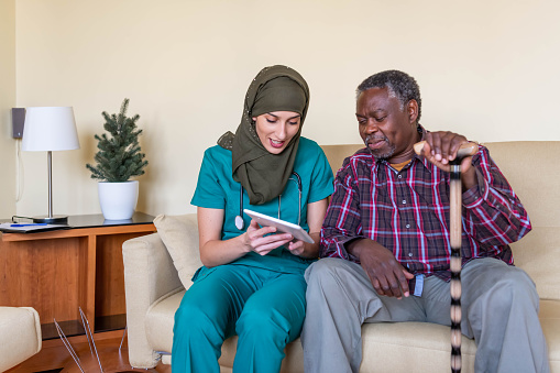 Arabian female doctor is at an in-home visit of her older patient of Afro-American ethnicity. A Muslim female healthcare worker in green uniform is writing a prescription to her Afro-American patient using her digital tablet.