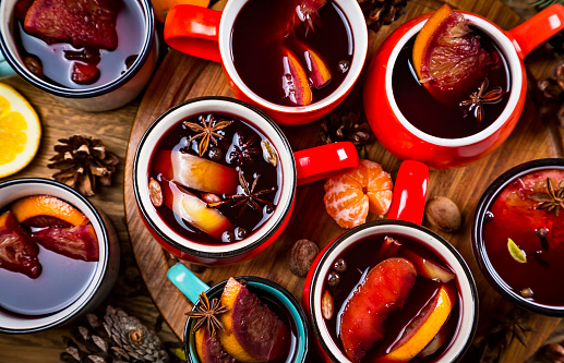 Top view of many colorful cups of traditional Christmas drink mulled red spiced wine with cinnamon stick, apples and citrus on wooden table with star anise and christmas fir tree. Christmas party.