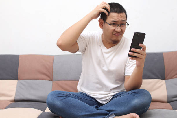 istock Confused Asian man scratch his head when looking at his phone while sitting on sofa, bad news 1287983215