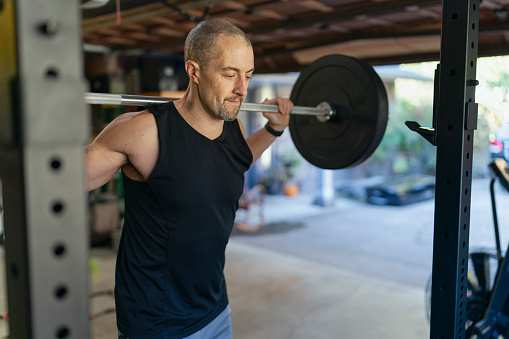 An athletic man doing a weight lifting workout routine uses a barbell to do back squats. The man is exercising in the home gym he set up in his garage while isolating at home during the Covid-19 pandemic.