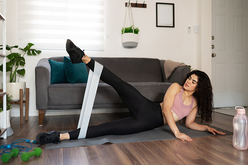 Sporty young woman doing leg raises using a resistance band. Latin woman working out at home