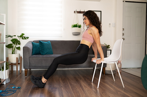 Attractive young woman exercising in her home. Sporty woman in her 20s doing tricep dips using a chair in the living room