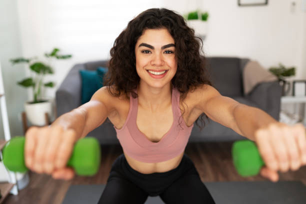 istock Attractive young woman squatting with two small weights at home 1288000755