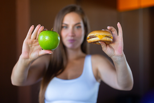 Young attractive girl is making choice between healthy and harmful food holding a green apple and a burger in her hands. Concept of proper nutrition, healthy food, fast food.