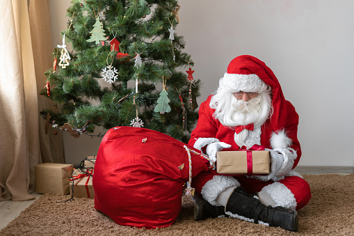 santa claus sits on the floor near the christmas tree untied his bag and took out a gift. box with red ribbon in Santa's hands.