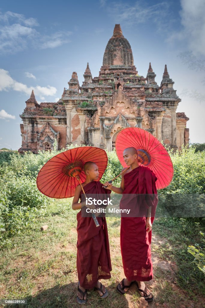 Novice monks standing together in front of ancient temple in Bagan Myanmar Happy young burmese buddhist novice monks in their typical red traditional religious veils, holding red sunshades, talking with each other standing together side beside in front of ancient Temple in Bagan. Bagan, Mandalay Region, Myanmar, Southeast Asia. Myanmar Stock Photo