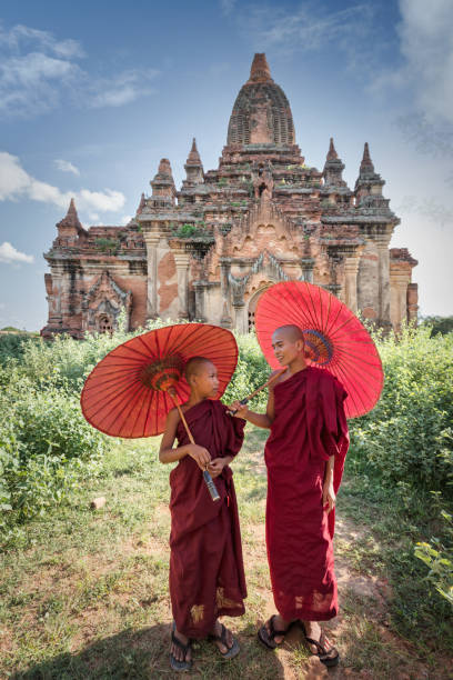 Novice monks standing together in front of ancient temple in Bagan Myanmar Happy young burmese buddhist novice monks in their typical red traditional religious veils, holding red sunshades, talking with each other standing together side beside in front of ancient Temple in Bagan. Bagan, Mandalay Region, Myanmar, Southeast Asia. bagan archaeological zone stock pictures, royalty-free photos & images