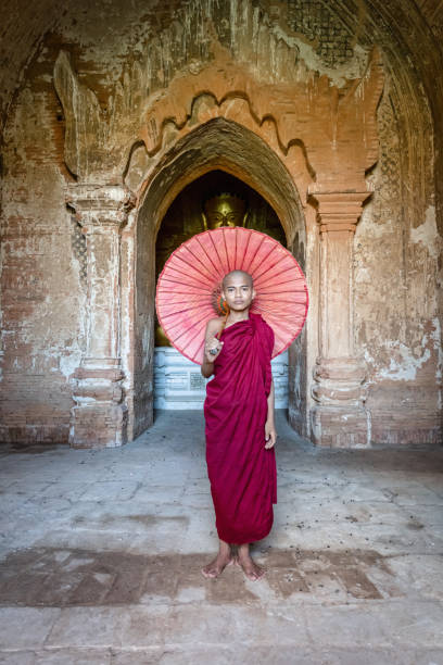 Novice Monk in front of Buddha Statue inside Buddhist Temple of Myanmar Burmese buddhist Novice Monk dressed in the buddhist traditional red religious dress and veil, holding a typical burmese red parasol standing straight in front of golden Buddha Statue inside ancient Temple in Bagan. Real Novice Monk Portrait. Bagan, Mandalay Region, Myanmar, Southeast Asia. bagan archaeological zone stock pictures, royalty-free photos & images