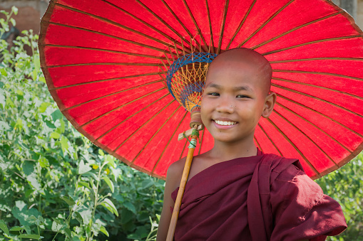 Bright and happy smiling burmese buddhist Novice Monk Child dressed in the buddhist traditional red religious dress and veil, holding a typical burmese red parasol. Real Novice Monk Children Portrait. Bagan, Mandalay Region, Myanmar, Southeast Asia.