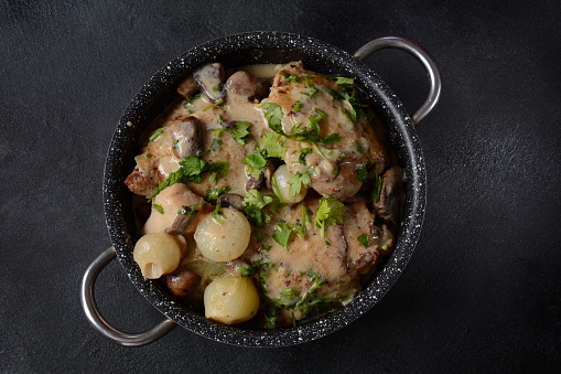 Fricassee - French Cuisine. Chicken stewed in a creamy sauce with mushrooms in casserole pan on a black table