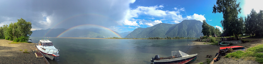 Panoramic view on double rainbow above river with mountains and cloudy sky on background.