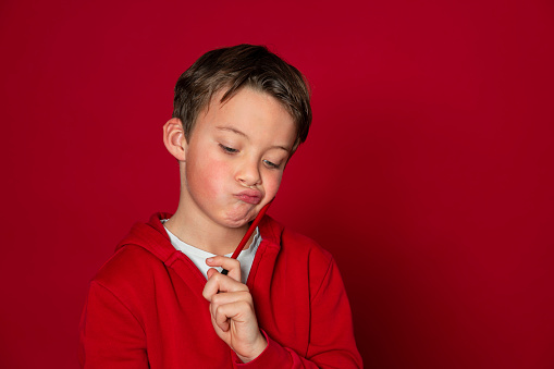 cool young school boy is posing with red pencil in ront of red background
