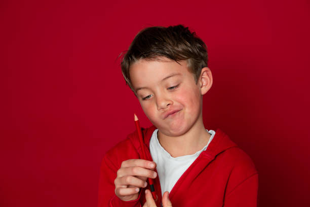 cool young school boy is posing with red pencil in ront of red background