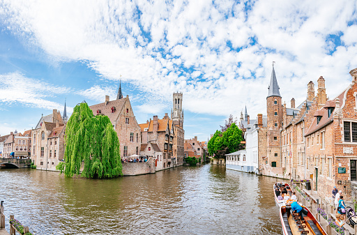 Sunny spring day in the historic city centre of Bruges - people are waiting for a boat tour on the embankment, surrounded by old medieval buildings. The Church of Our Lady and Belfry Tower is on the background.