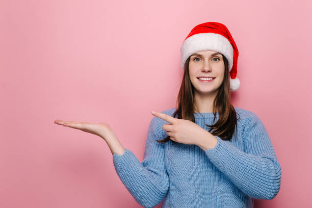 istock Attractive young girl keeps palm raised, points with forefinger against pink wall for advertisement, dressed in Christmas red hat and winter blue sweater. Happy New Year celebration merry holiday 1288067948
