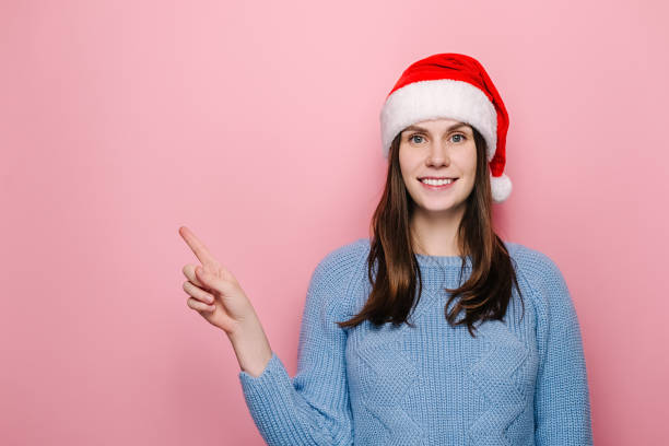 istock Lovely young woman in Christmas red hat points fore finger right, suggests going this direction, demonstrates awesome product, wears winter sweater, isolated on pink background, has positive smile 1288067949