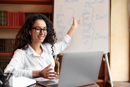 Distance Education And Learning Concept. Portrait of smiling female English teacher sitting at desk and pointing at whiteboard, explaining rules to students. Excited lady wearing glasses and earphones