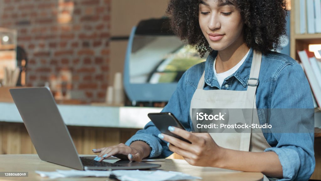 Owner of coffee business works in accounting with social media Owner of coffee business works in accounting with social media. Young african american serious woman in apron looking at smartphone and typing on laptop in interior of modern coffee shop, panorama Owner Stock Photo