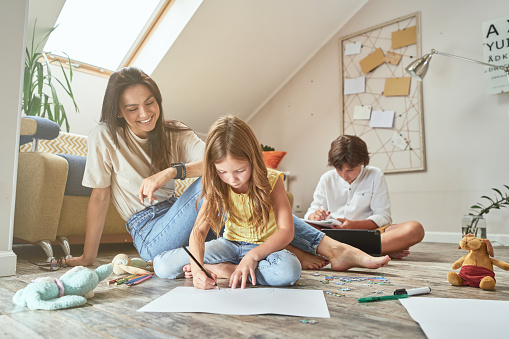 Spending time with children at home. Little girl sitting together with mother on the floor in living room and drawing while her brother doing homework on the background. Family concept