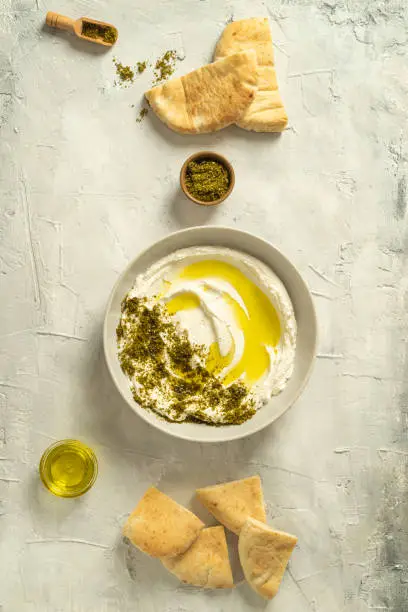 Photo of Popular middle eastern appetizer labneh or labaneh, soft white goat milk cheese with olive oil, hyssop or zaatar, served with pita bread on a grey table, top view,