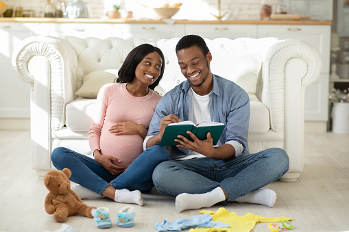 Happy black couple making checklist of baby things for maternity hospital at home. Loving African American husband and wife getting ready for child delivering, preparing stuff for clinic