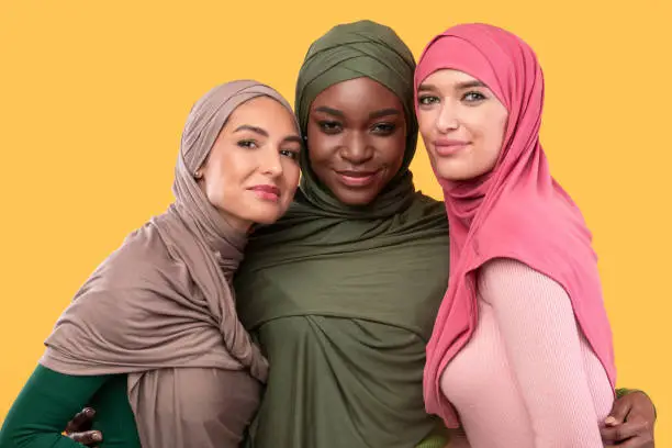 Three Muslim Women In Hijab Headscarf Embracing Posing Smiling To Camera Standing Together In Studio On Yellow Background. Modern Islamic Ladies Portrait. Arabic Female Fashion Concept