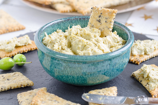 Delicious fresh homemade artichoke dip in a turquoise bowl with sesame crackers, a creamy ans tangy sauce made with artichoke hearts, garlic, capers, olive oil,green olives and lemon juice