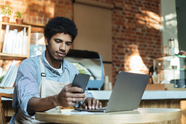 Social networking, devices and small business during covid-19 pandemic Social networking, devices and small business during covid-19 pandemic. Busy african american man in apron sits at table and works in smartphone and laptop in interior of loft coffee shop, copy space busy restaurant tables stock pictures, royalty-free photos & images