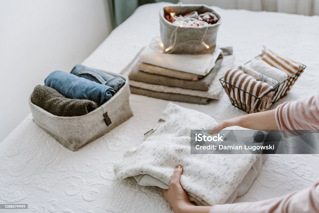 Woman folding stack of fresh laundry, organizing clothes and towels in boxes and baskets. Woman folding clothes in bedroom, organizing laundry in boxes and baskets. Concept of minimalism lifestyle and japanese t shirt folding system. Tidy up wardrobe Organization Stock Photo