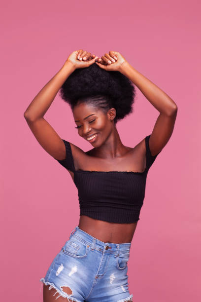 splendida giovane ragazza afroamericana con i capelli afro in posa con le braccia alzate vestite con top nero e jeans in denim su sfondo rosa sporco - arms lifted foto e immagini stock
