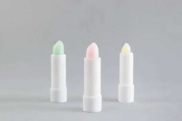 Photo of White tube of pink, green and yellow lip balm to protect and nourish lips on a gray background. Winter season cosmetics. Container layout. Gift idea for a woman. Copy space