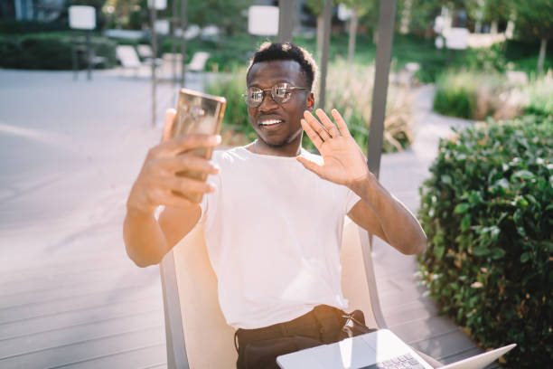 sonriente de piel oscura blogger macho que dispara contenido en la cámara del teléfono inteligente para vlog sentado en la terraza del café, feliz estudiante hombre americano africano haciendo videollamada a través del teléfono móvil y conexión 4g - correspondence waving horizontal outdoors fotografías e imágenes de stock