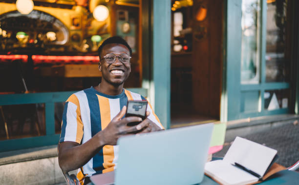 hombre negro alegre con teléfono inteligente y portátil en la cafetería de la calle en la terraza de verano - hipster people surfing the net internet fotografías e imágenes de stock