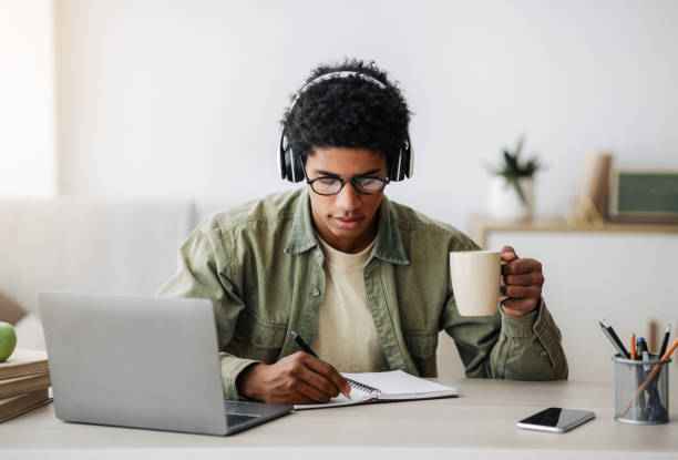 istock Busy teen guy drinking coffee during online lesson on laptop, writing down info in notebook, indoors 1288089729
