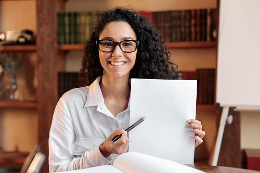 Sign Here. Young smiling confident woman wearing eyeglasses pointing to a blank empty sheet of paper, holding a pen, looking and showing it at camera. Presentation, Agreement, Business Concept.