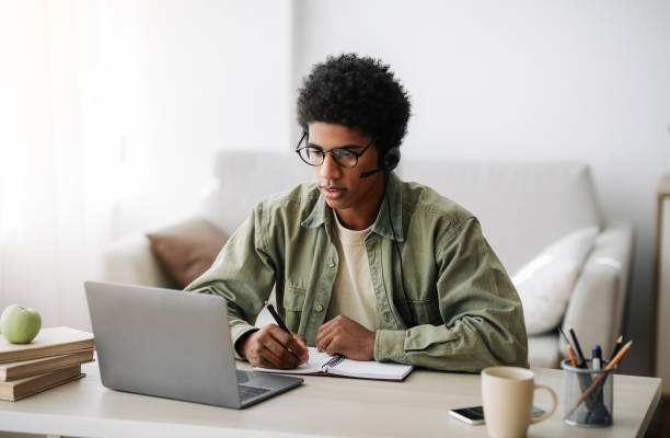 istock Industrious black teenager in headset studying remotely from home, writing in notebook 1288090024