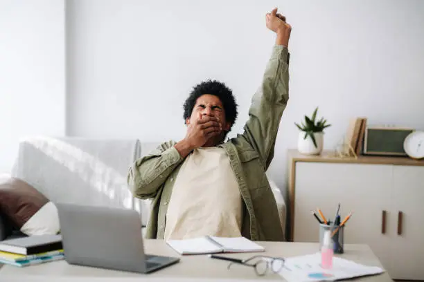 Tired black student yawning and stretching during his remote studies from home. African American youth exhausted from getting ready for test or writing coursework, feeling sleepy in front of laptop