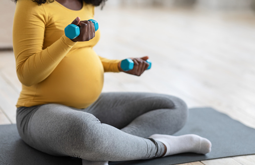 Unrecognizable pregnant african american woman exercising with dumbbells at home, sitting on fitness mat, copy space. Cropped of expecting black lady lifting small barbells up, healthy lifestyle