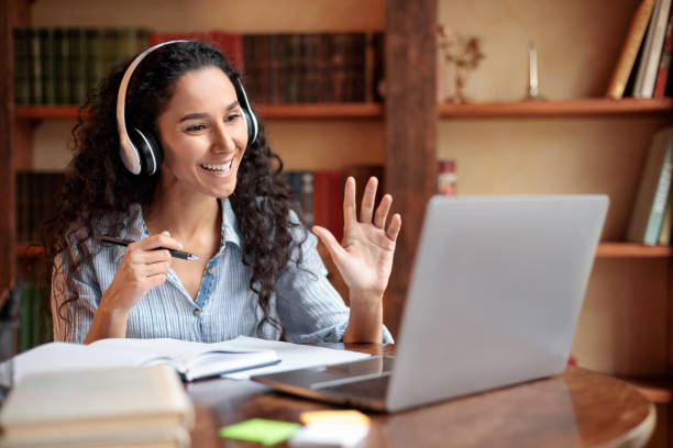 Woman sitting at desk, using computer and waving to webcam Teleconference And Telecommuting. Portrait of smiling woman sitting at desk, having video call on laptop, waving to webcam. Distance Education. Positive lady wearing headest at virtual meeting online tutors stock pictures, royalty-free photos & images