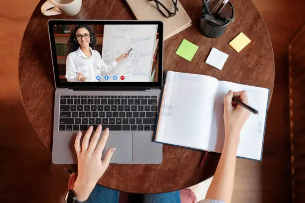 Distance Education Concept. Above top view of female student sitting at desk, writing in notebook, taking notes, using laptop, having online class with teacher. Remote studying from home, quarantine