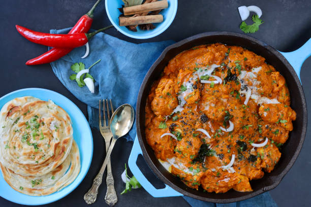 Stock photo showing elevated surface view of turquoise blue pan of chicken tikka curry sauce. Butter chicken breast chunks sprinkled with red onion and mustard seeds, served with lachha paratha (layered flatbread). imagen de la sartén azul turquesa llena de pollo de mantequilla / curry tikka, grandes trozos de carne de pechuga de pollo en salsa de curry con guarnición de rodajas de cebolla roja y semillas de mostaza, lachha paratha (pan plano en capas), vista eleva