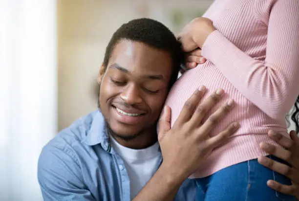 Photo of Young black man pressing his ear against his wife's pregnant belly, listening to his baby's heartbeat indoors