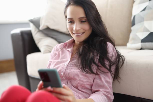 istock Woman sit on floor near sofa, look at screen and smile. 1288094059