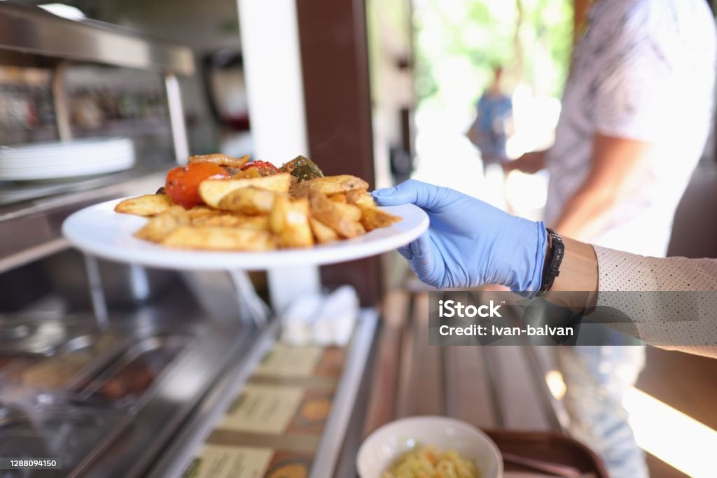 Female hand in blue medical glove hold white plate with fried potatoes and stewed vegetables. Female hand in blue medical glove hold white plate with fried potatoes and stewed vegetables. Self service restaurant with tray. Cafeteria Stock Photo