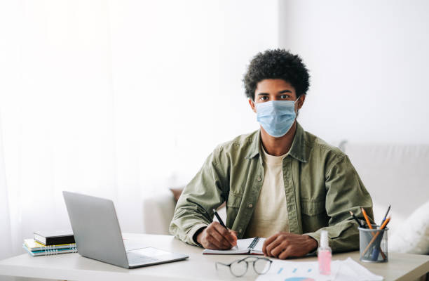 istock Web based education during covid-19. Black teen guy in surgical mask taking notes during online lecture on laptop 1288094527