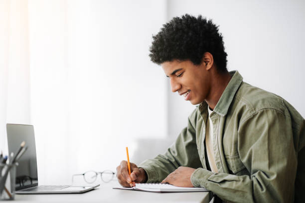 istock Web-based training concept. Black male student writing in notebook in front of laptop at home, empty space 1288095614