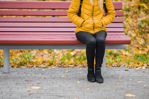 One young girl in yellow jacket and black trousers sitting on wooden bench at park in autumn day. Front view. Closeup.