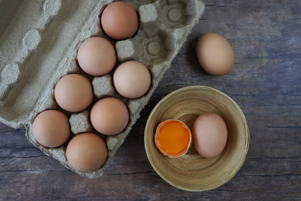 Stock photo showing close-up view of batch of brown chicken eggs in an open disposable cardboard egg box on wooden table top beside bowl containing eggs with one broken showing yolk.