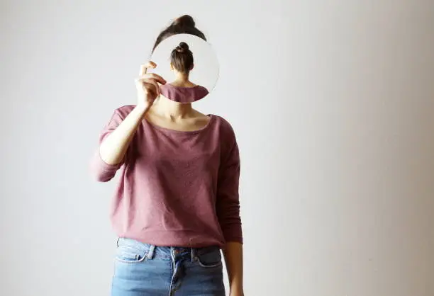 Young woman holding a mirror in front of her face. Faceless, identity not recognizable, the mirror is showing her backhead. Hiding her face.