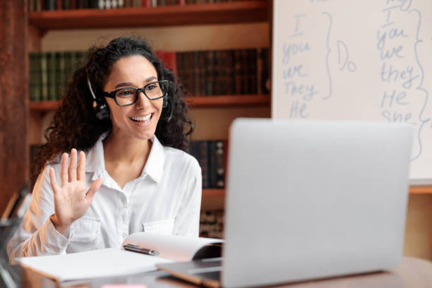 Lady sitting at desk, using computer and waving to webcam Distance Education. Positive lady wearing glasses and wireless headest at virtual meeting, sitting at desk, having video call on laptop, waving to webcam. Woman studying or teaching online at home teaching stock pictures, royalty-free photos & images